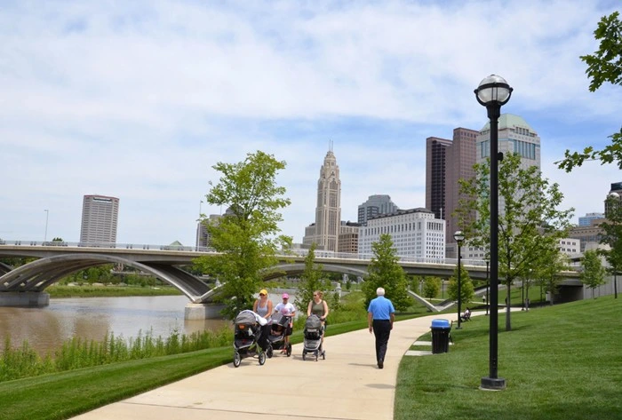 Scioto Mile riverfront walkway with people strolling beside the Columbus skyline and bridges in downtown Columbus.