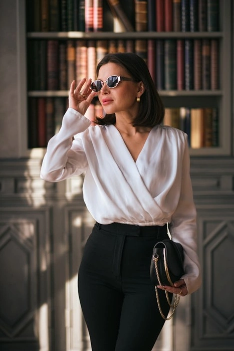 Smart casual dress for women styled with a wrap blouse, tailored trousers, and sunglasses in front of a bookshelf