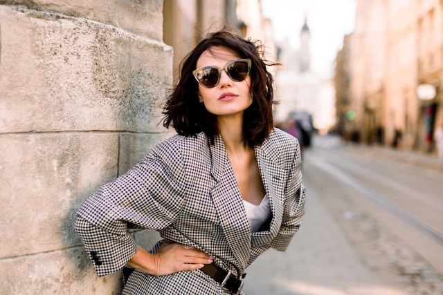 Woman leaning against a stone wall wearing a plaid blazer with sunglasses for a smart street style outfit