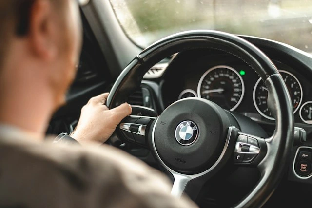 Person's hand on a BMW steering wheel while driving, dashboard gauges visible