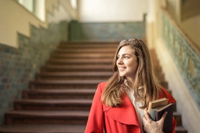 Smiling young professional in a red blazer climbing stairs with a book, symbolizing career growth