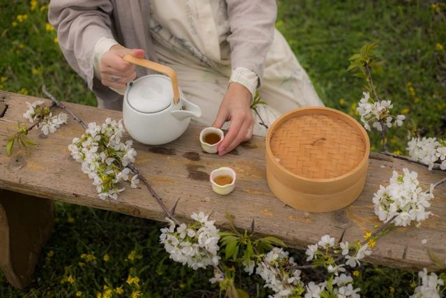 Person pouring tea from a compact ceramic teapot into small cups on a wooden bench surrounded by cherry blossoms with a woven storage set nearby