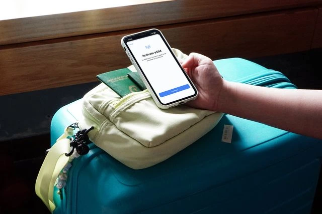 Traveler activating an eSIM in Vietnam on a smartphone, resting on a passport, a pale green crossbody bag and a turquoise suitcase