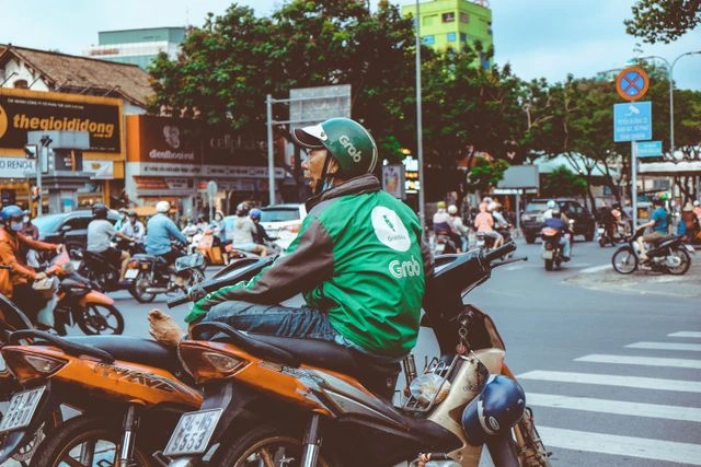 Grab motorbike driver in a green jacket and helmet waiting at a busy street intersection in Vietnam, surrounded by scooters and shopfronts Travelers queuing at Gate 14 in a Vietnam airport as a flight attendant in a green uniform greets passengers before boarding Two friends filming a video on a phone at an outdoor event, one wearing a Vietnam flag t-shirt, sharing travel moments online Winding mountain road through green terraced hillsides in northern Vietnam, dotted with small houses and haystacks, where travel signal can be patchy