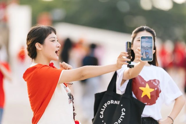 Two friends filming a video on a phone at an outdoor event, one wearing a Vietnam flag t-shirt, sharing travel moments online