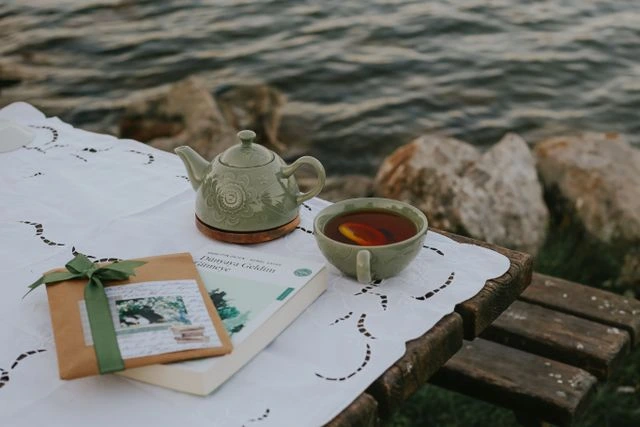 Small green teapot and matching teacup on an embroidered tablecloth by the water with a book and gift-wrapped package nearby