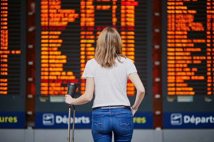 A traveler holding a suitcase and a coffee cup stands in front of an orange departures board, checking whether her flight is delayed or cancelled