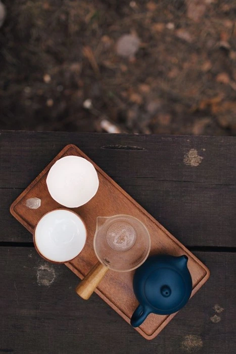Overhead view of a tea set for camping with white cups, a glass fairness pitcher, and a navy teapot arranged on a wooden tray