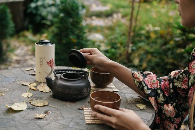 Person lifting the lid of a travel-friendly cast iron teapot beside wooden teacups on a stone table surrounded by fallen leaves