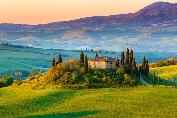 Villa vs. hotel in Tuscany — stone farmhouse surrounded by cypress trees on a green hillside at sunset
