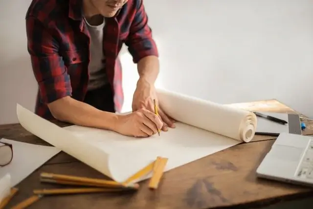 Contractor in a plaid shirt sketching project plans on a large paper roll before preparing the job’s invoicing