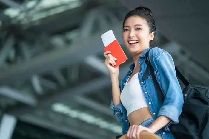 Smiling traveler in a denim jacket holding a red passport and an onward ticket at the airport, ready for her visa check