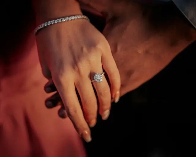Close-up of a pear-shaped halo diamond engagement ring on a woman’s hand as the couple holds hands, a design to compare when choosing a ring long distance