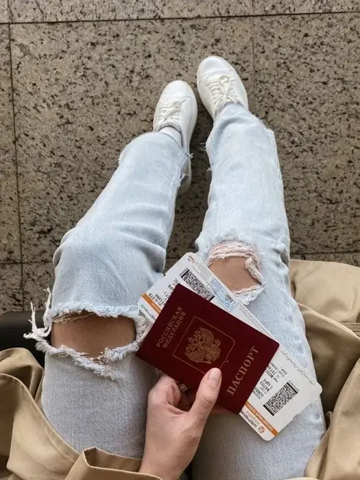 Traveler in ripped jeans and white sneakers holding a burgundy passport and printed boarding passes, waiting with her onward ticket for a visa or check-in review