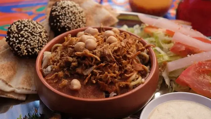 Bowl of Egyptian koshari topped with crispy fried onions and chickpeas, served with sesame falafel, pita bread and fresh salad