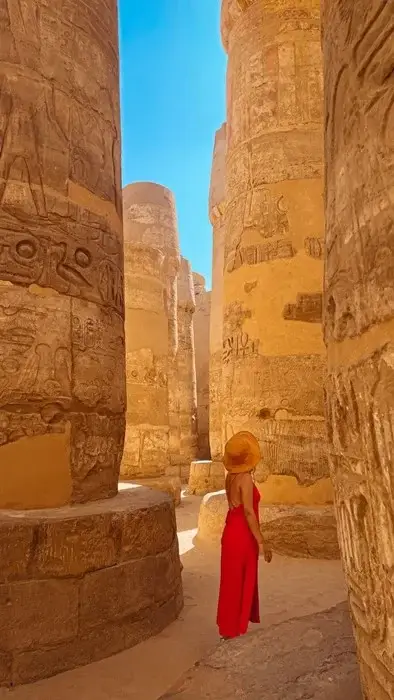 Woman in a red dress and straw hat standing among the towering carved columns of the Hypostyle Hall at Karnak Temple in Luxor