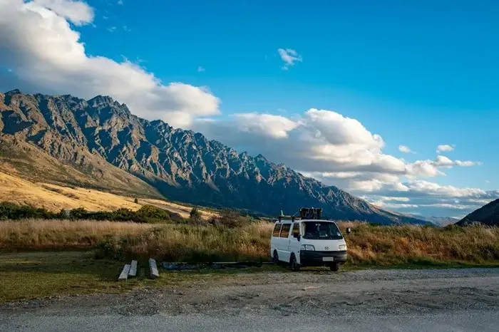 A small white campervan parked at a gravel roadside stop below a jagged New Zealand mountain range, showing the kind of overnight spot that affects trip pricing