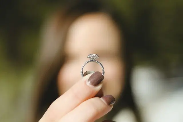 Woman holding up a round halo diamond ring with a pavé band toward the camera, checking the setting before the couple decides on the final engagement ring