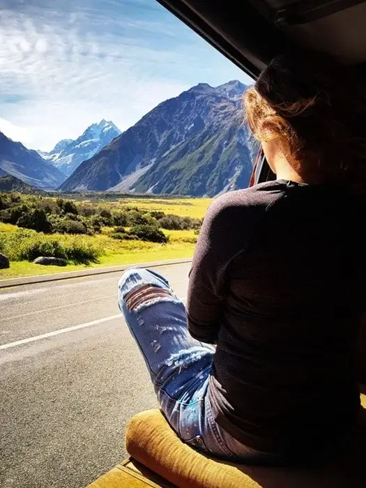 A traveler sits in the open doorway of a campervan, looking out at a snow-capped peak and green valley beside a quiet New Zealand road