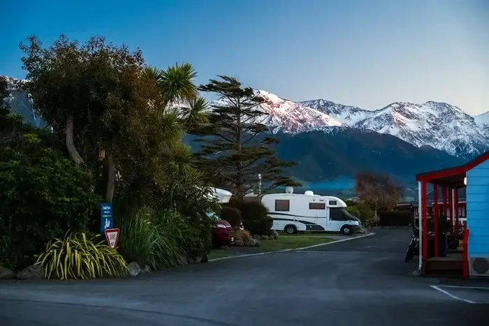 A motorhome parked at a New Zealand holiday park at dusk, with snow-capped mountains glowing pink behind a blue cabin, showing a paid campsite option when pricing overnight stops