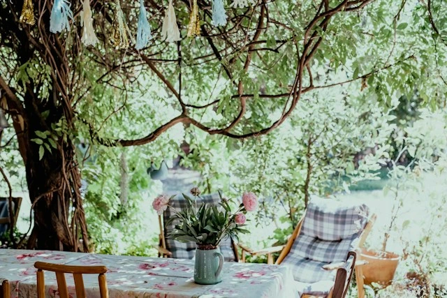 Outdoor dining table set under a shady tree at home, with a floral tablecloth, a jug of pink peonies, plaid-cushioned chairs, and a paper tassel garland overhead