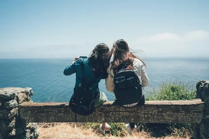 Two hikers with backpacks sitting on a wooden railing at a Big Sur overlook, scanning the Pacific Ocean for wildlife