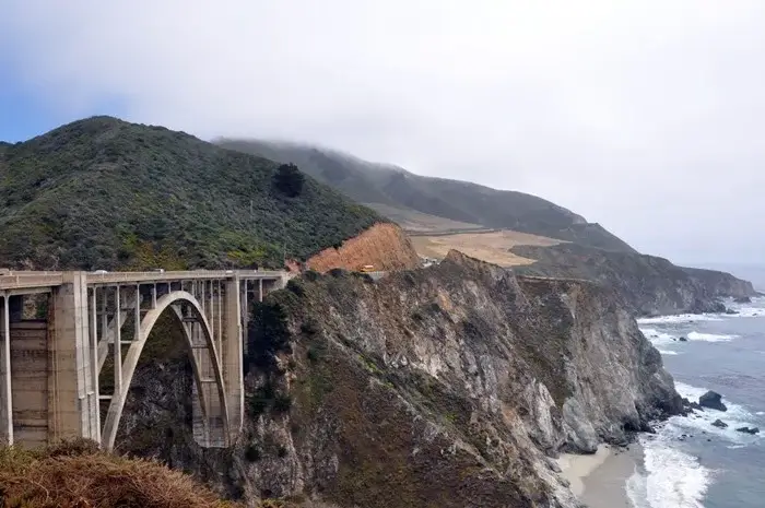 Bixby Creek Bridge arching over a rugged canyon as fog rolls across the green hills and rocky Big Sur coastline