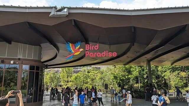 Visitors and families arrive at the Bird Paradise entrance in Mandai, with the park's colorful sign beneath a large curved canopy