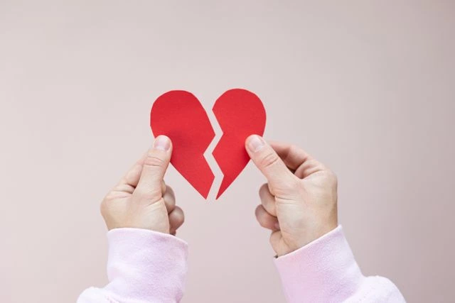 Hands in pink sleeves holding two halves of a red paper heart, a simple symbol of a breakup against a soft blush background