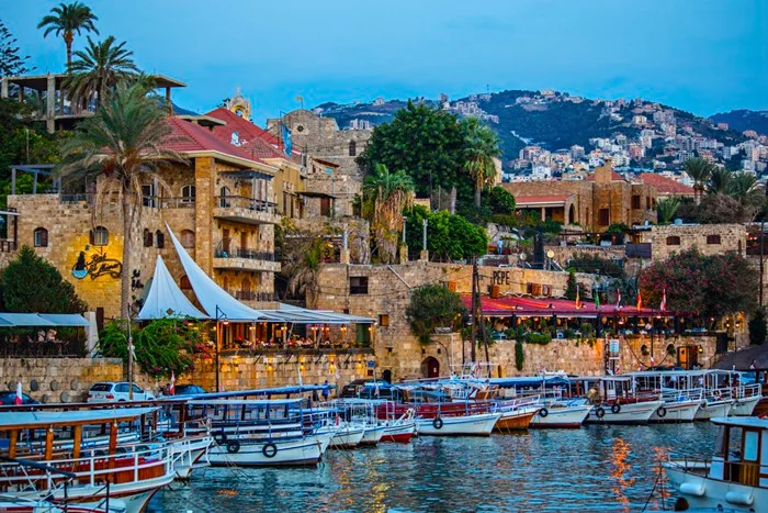 Fishing boats moored in the ancient port of Byblos at dusk, with sandstone buildings, palm trees, and hillside homes, a highlight for Lebanon travel