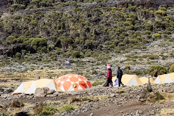 Tents pitched at a rocky high camp while climbing Kilimanjaro, with a porter carrying a bucket on her head