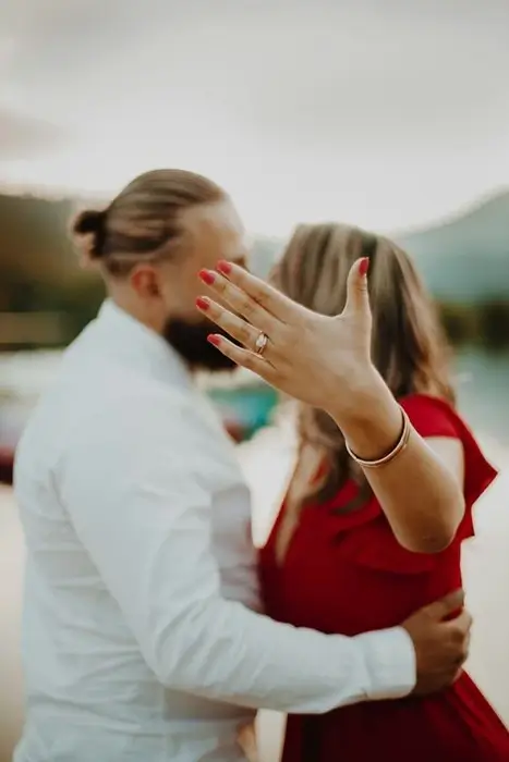 Woman in a red dress holding out her hand to show her engagement ring while hugging her partner by a lake, celebrating after choosing an engagement ring for LDR couple shopping