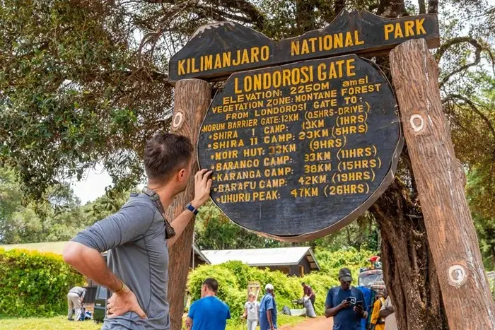 Climbing Kilimanjaro guide: hiker reading the Londorosi Gate sign with distances to each camp and Uhuru Peak