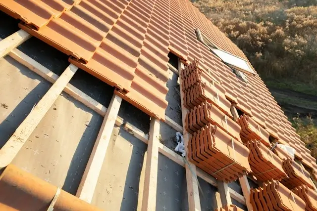Clay tile roof installation in progress, with stacked orange tiles on wooden battens over underlayment
