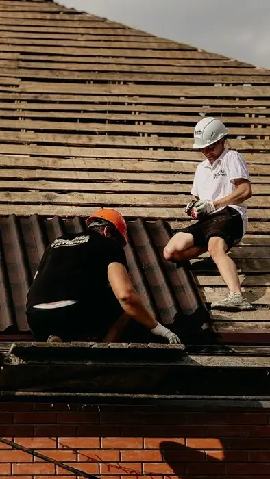 Two roofers in hard hats laying dark roof tiles over exposed wooden battens on a brick building