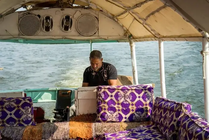 Local boatman steering a small motorboat on the Nile with purple patterned cushions under a shaded canopy