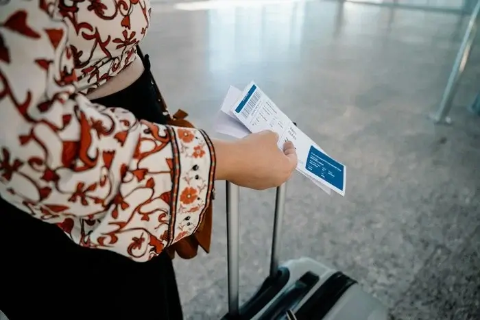 Traveler in a floral patterned top holding printed flight tickets beside a rolling suitcase in an airport terminal, with her onward ticket ready for the airline service desk