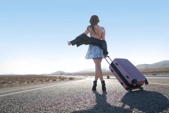 Woman in a sequin dress pulling a pink suitcase and hitchhiking on a desert road, ready to travel and start fresh after a breakup