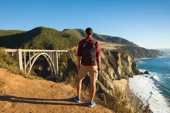 Hiker in a plaid shirt and backpack standing on a sunny bluff overlooking Bixby Creek Bridge, watching the coast for wildlife