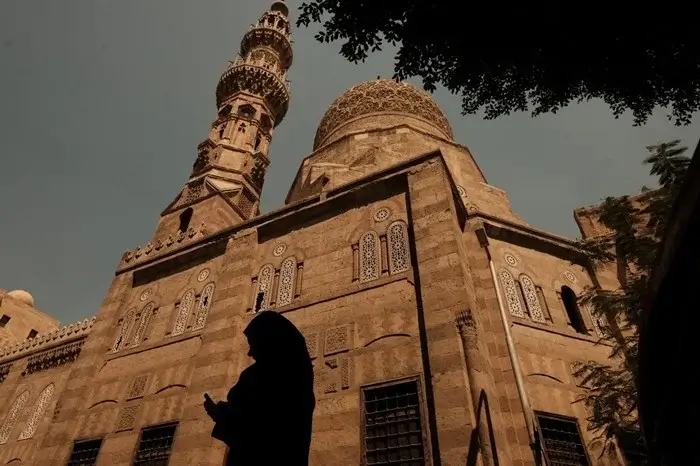 Low-angle view of a Mamluk-era mosque in historic Cairo, its intricately carved stone dome and minaret rising above a silhouetted figure