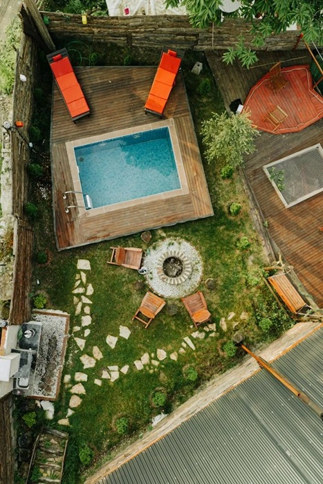 Aerial view of a fenced backyard garden with a small pool on a wooden deck, orange sun loungers, a stone fire pit circled by wooden chairs, and a stepping-stone path