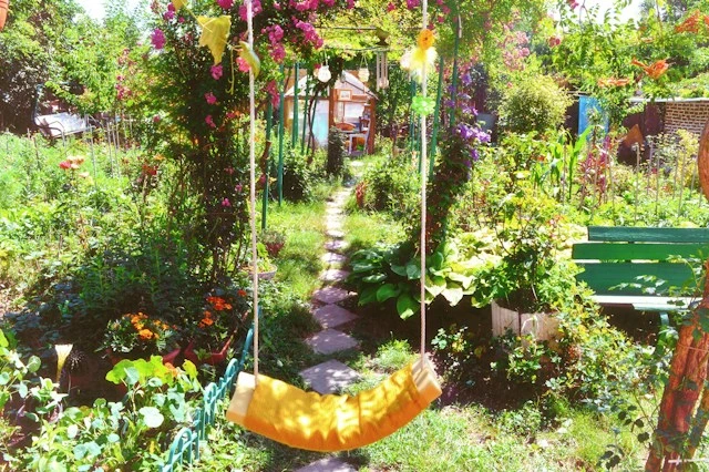 Sunny home garden with a yellow rope swing, a stepping-stone path leading to a small shed, climbing pink roses, lush flower beds, and a green wooden bench