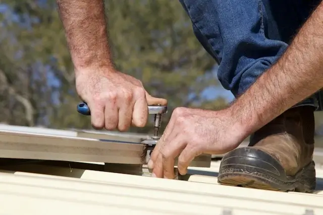 Two roofers in hard hats laying dark roof tiles over exposed wooden battens on a brick building