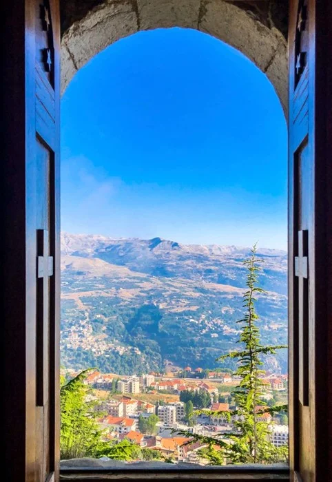 Lebanon travel guide view of red-roofed town and mountains through an arched stone window with wooden shutters