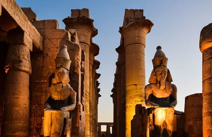 Seated statues of Ramesses II and towering columns lit up at dusk at Luxor Temple in Egypt