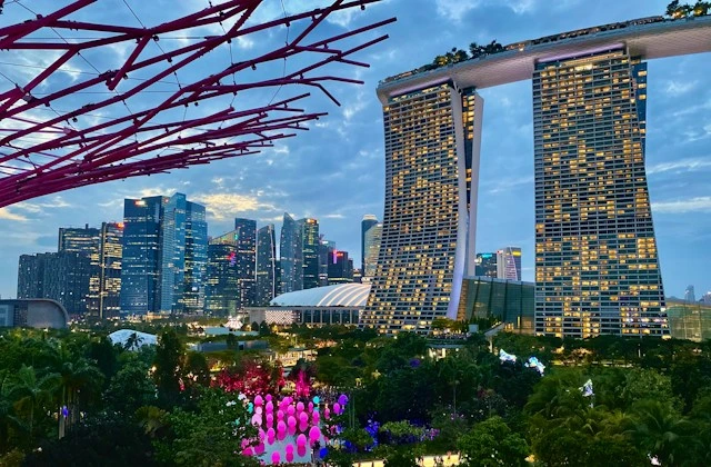 Marina Bay Sands and the city skyline at dusk, seen from Gardens by the Bay, with a Supertree's red branches framing the view and colorful lights glowing in the gardens below