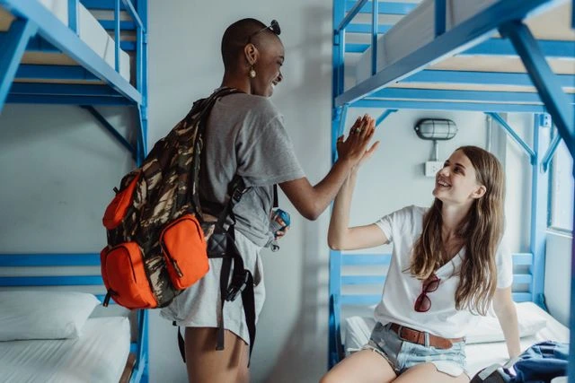 Two smiling women sharing a high five in a hostel dorm with blue bunk beds, showing how solo travel makes it easy to meet new friends