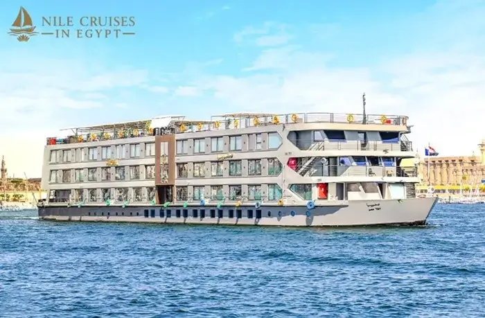 Modern 4-deck cruise ship sailing on the Nile with ancient temple columns lining the riverbank behind it