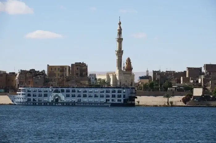 Nile river cruise ship docked along the riverbank beneath a tall mosque minaret in an Egyptian town