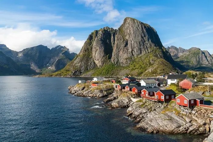 Red rorbuer fishing cabins on a rocky shoreline beneath a towering granite peak in Hamnøy, Lofoten Islands, Norway
